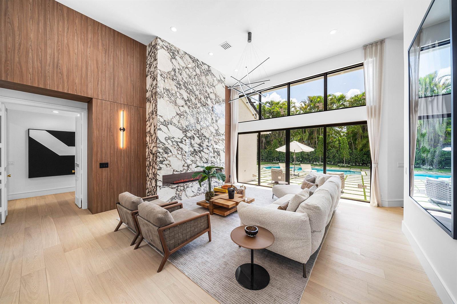 A modern living room features a marble fireplace, wood paneling, and floor-to-ceiling windows overlooking a pool — 7028 Mandarin Drive, Boca Raton FL 33433, BOCA RATON, FL