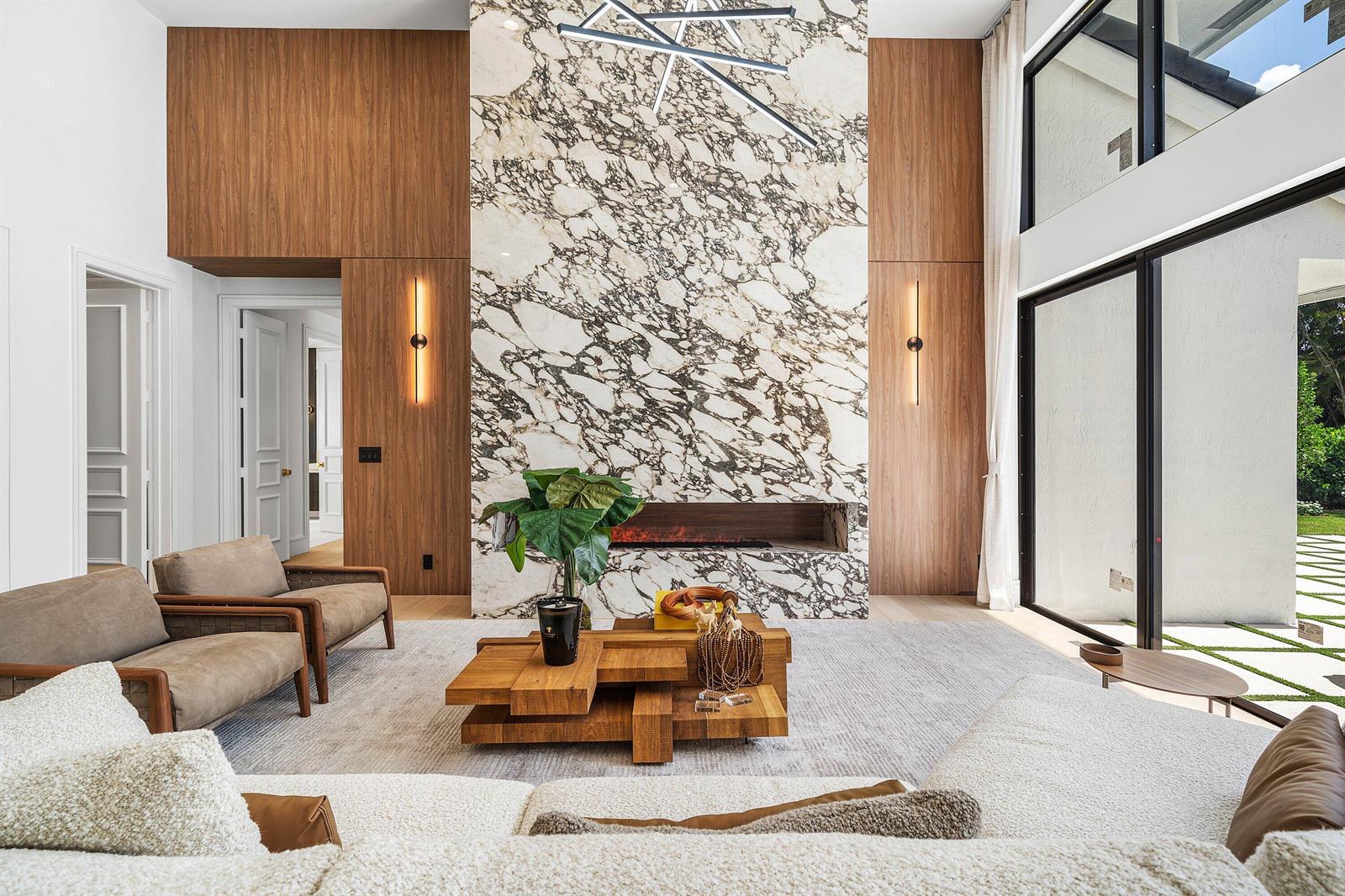 A modern living room features a large marble fireplace, wood paneling, and floor-to-ceiling windows — 7028 Mandarin Drive, Boca Raton FL 33433, BOCA RATON, FL