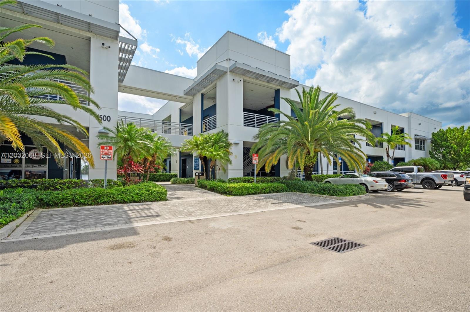 A modern two-story commercial building features white facades, palm trees, and a paved parking area — 1500 NW 89th Ct # 112, Doral FL 33172, DORAL, FL