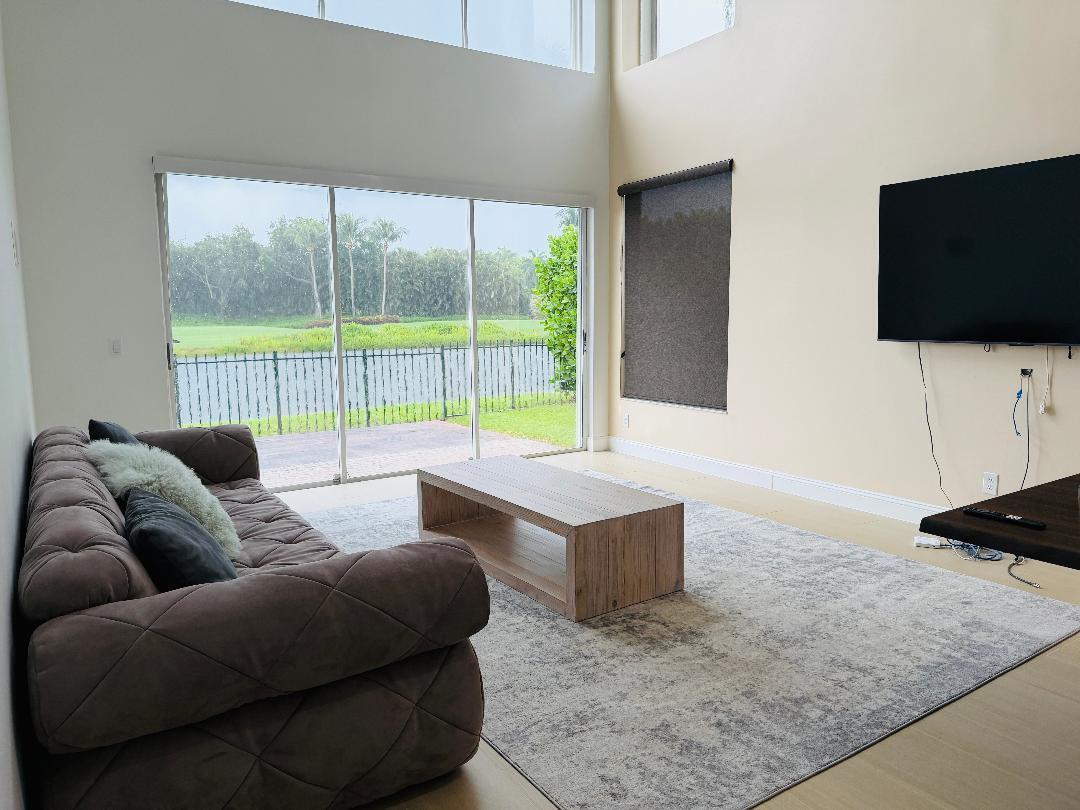 A living room with a brown sofa, wooden coffee table, and large windows overlooking a lake — 5855 NW 42nd Way, Boca Raton FL 33496, BOCA RATON, FL