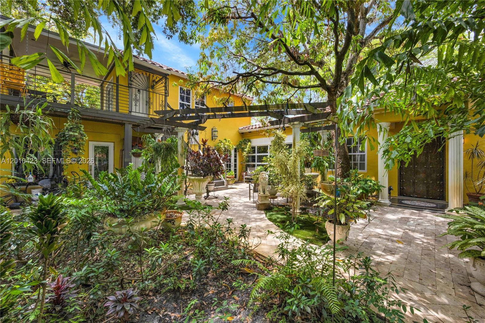 A yellow two-story house features a paved courtyard with a wooden pergola and lush landscaping — 650 NE 76th St, Miami FL 33138, MIAMI, FL