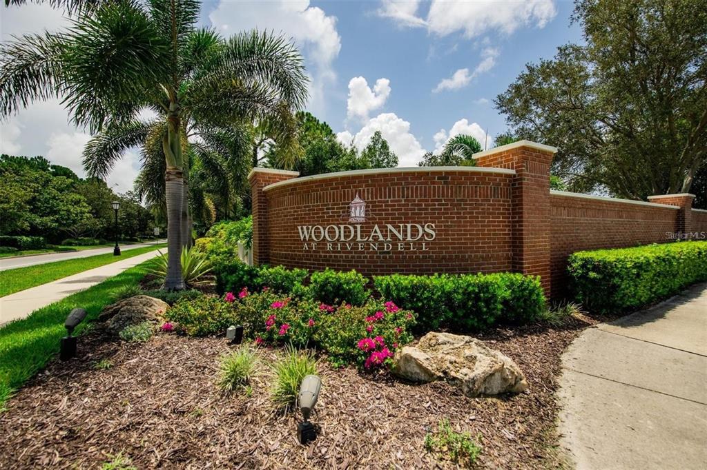 A brick entrance sign for Woodlands at Rivendell stands beside a sidewalk and palm tree — 1036 OAK MEADOW LANE, OSPREY, FL