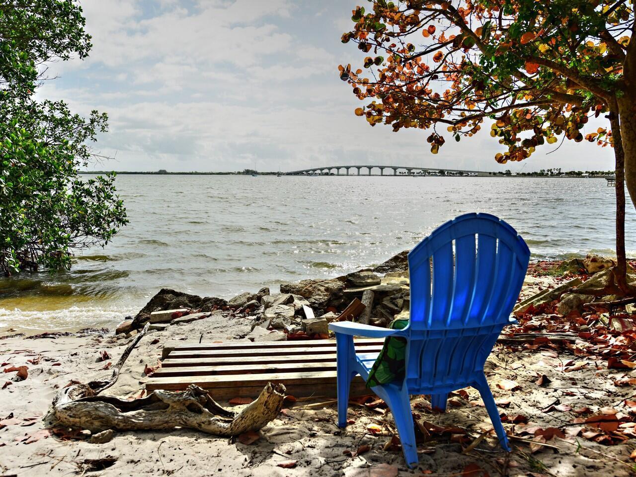 A blue Adirondack chair sits on a sandy shore overlooking a bay and a bridge — 4220 NE Indian River Dr. Drive # 2, Jensen Beach FL 34957, JENSEN BEACH, FL