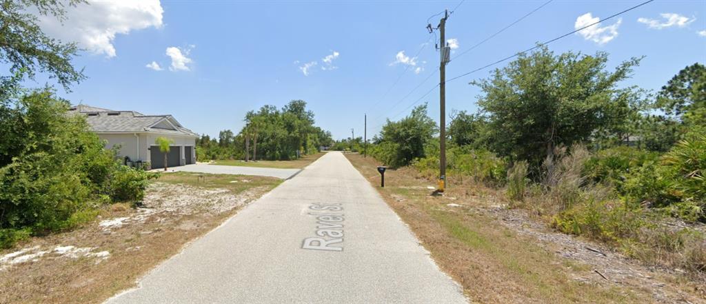 A paved street leads past a single-story house and undeveloped wooded lots under a blue sky — 9204 & 9196 RAVEL STREET, PORT CHARLOTTE, FL