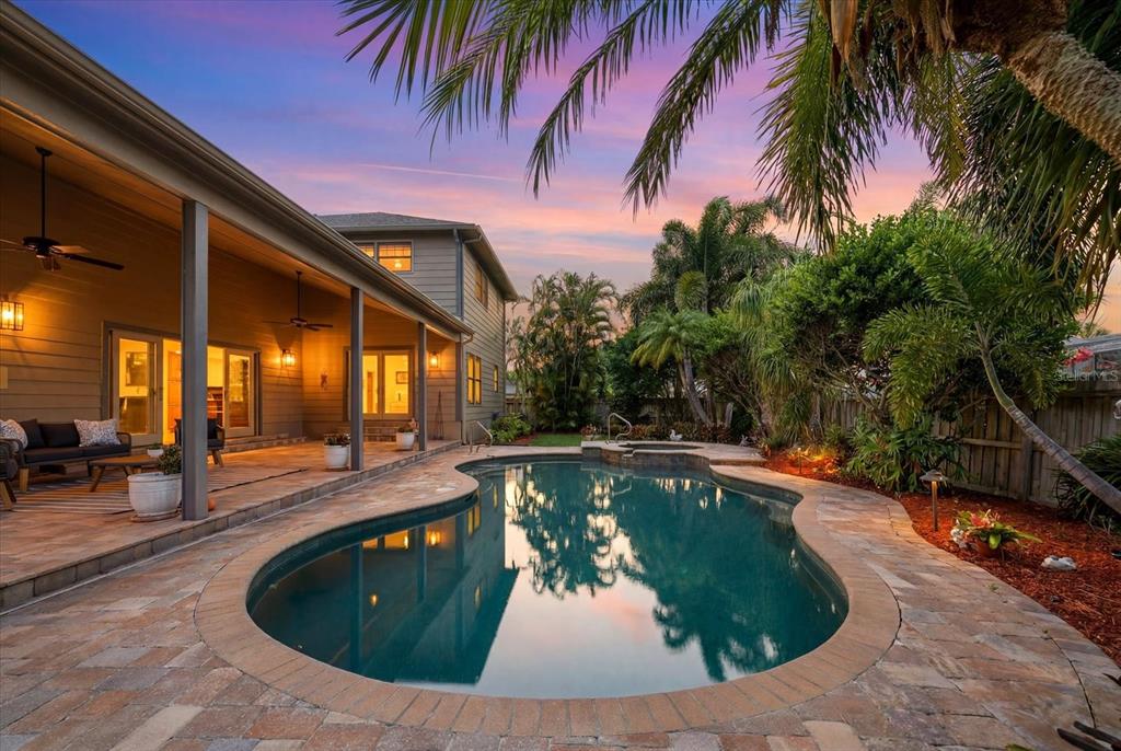 A backyard features a curved swimming pool, a stone patio, and a covered porch at sunset — 2917 SANDPIPER PLACE, CLEARWATER, FL