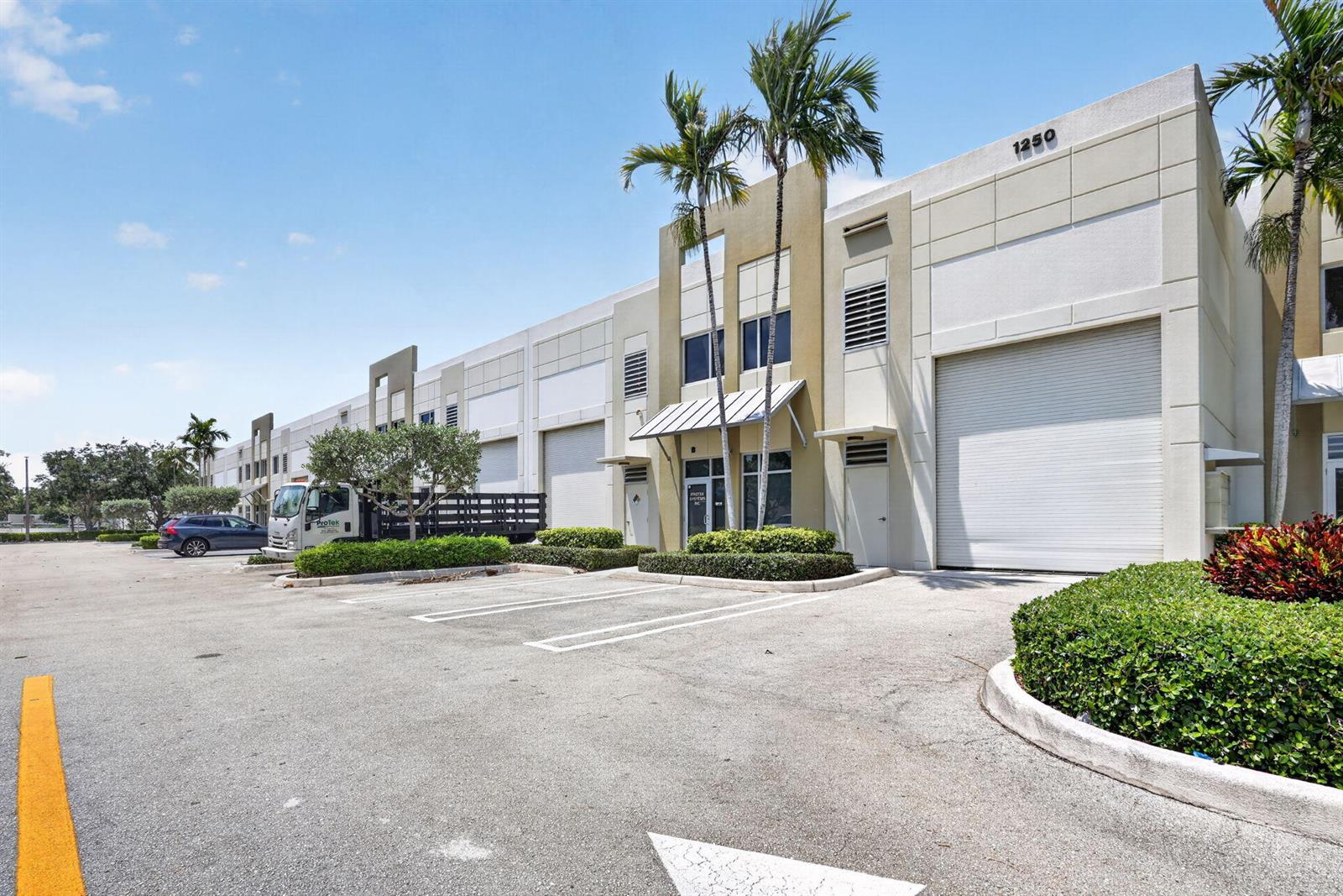 An exterior view of a commercial warehouse building with parking spaces and palm trees — 1250 Wallace Drive # A, Delray Beach FL 33444, DELRAY BEACH, FL