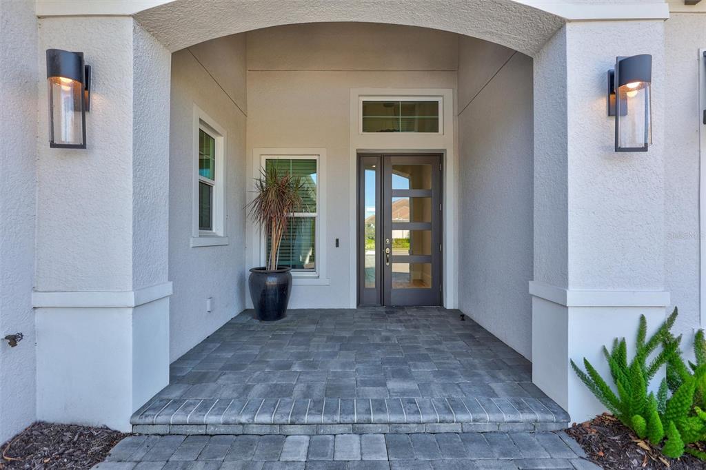 A covered entryway features a dark-framed glass door, stone paver flooring, and two wall-mounted lanterns — 8278 PAVIA WAY, BRADENTON, FL