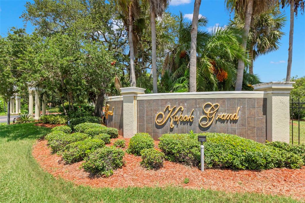 A stone monument sign for Kapok Grand stands in a landscaped garden with palm trees — 11117 KAPOK GRAND CIRCLE, MADEIRA BEACH, FL