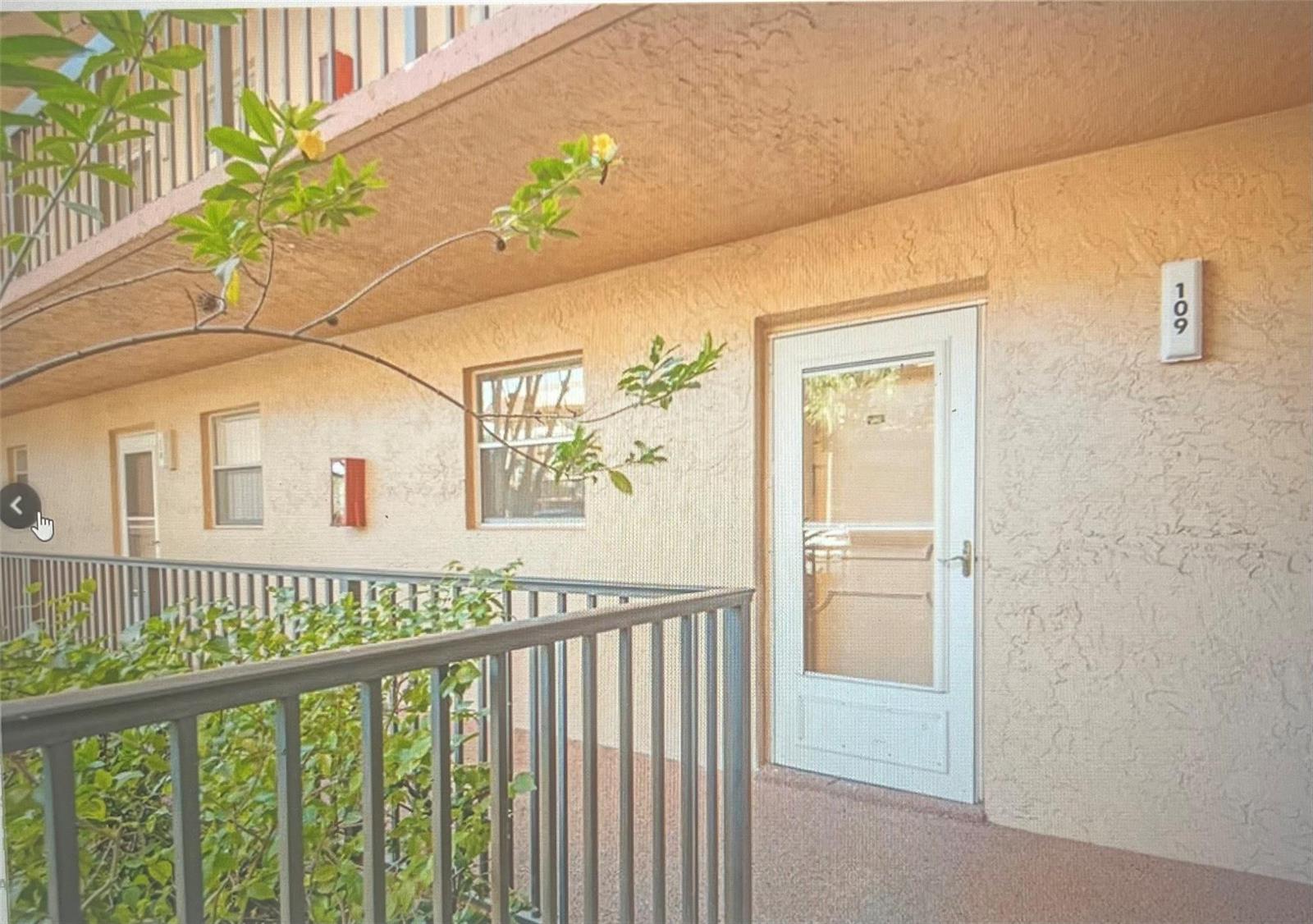An exterior view of a ground-floor apartment entrance with a white door and metal railing — 3140 Holiday Springs Boulevard # 109, Margate FL 33063, MARGATE, FL