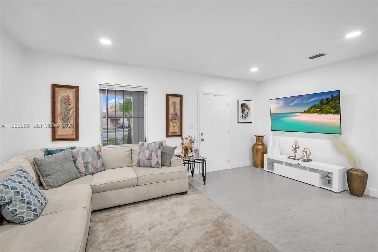 A living room features a beige sectional sofa, a wall-mounted television, and tiled flooring — 14140 NE 10th Ave, North Miami FL 33161, NORTH MIAMI, FL
