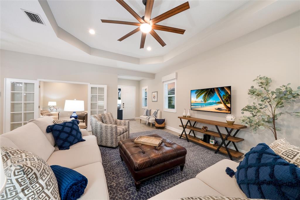 A living room features a tray ceiling, a ceiling fan, and a wall-mounted television — 12825 COASTAL BREEZE WAY, BRADENTON, FL
