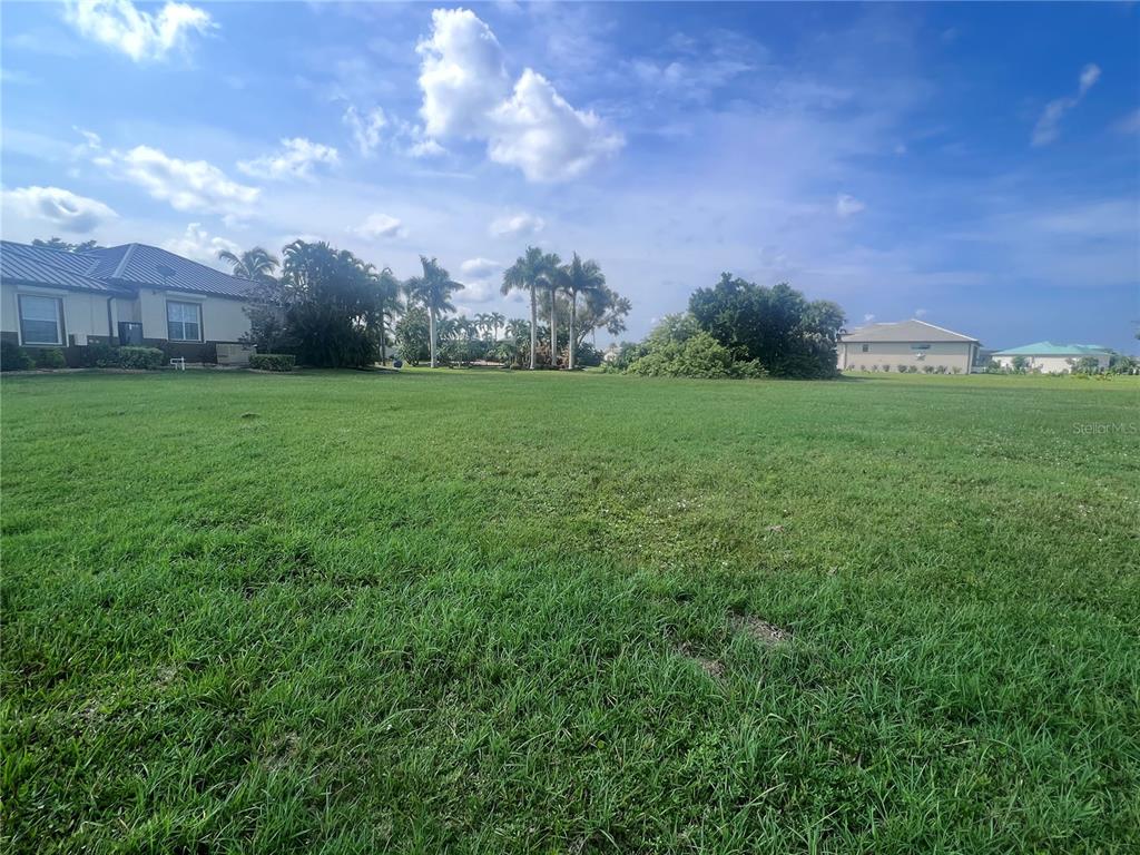 A large, grassy vacant lot sits in front of residential houses under a blue sky — 24183 SANTA INEZ ROAD, PUNTA GORDA, FL