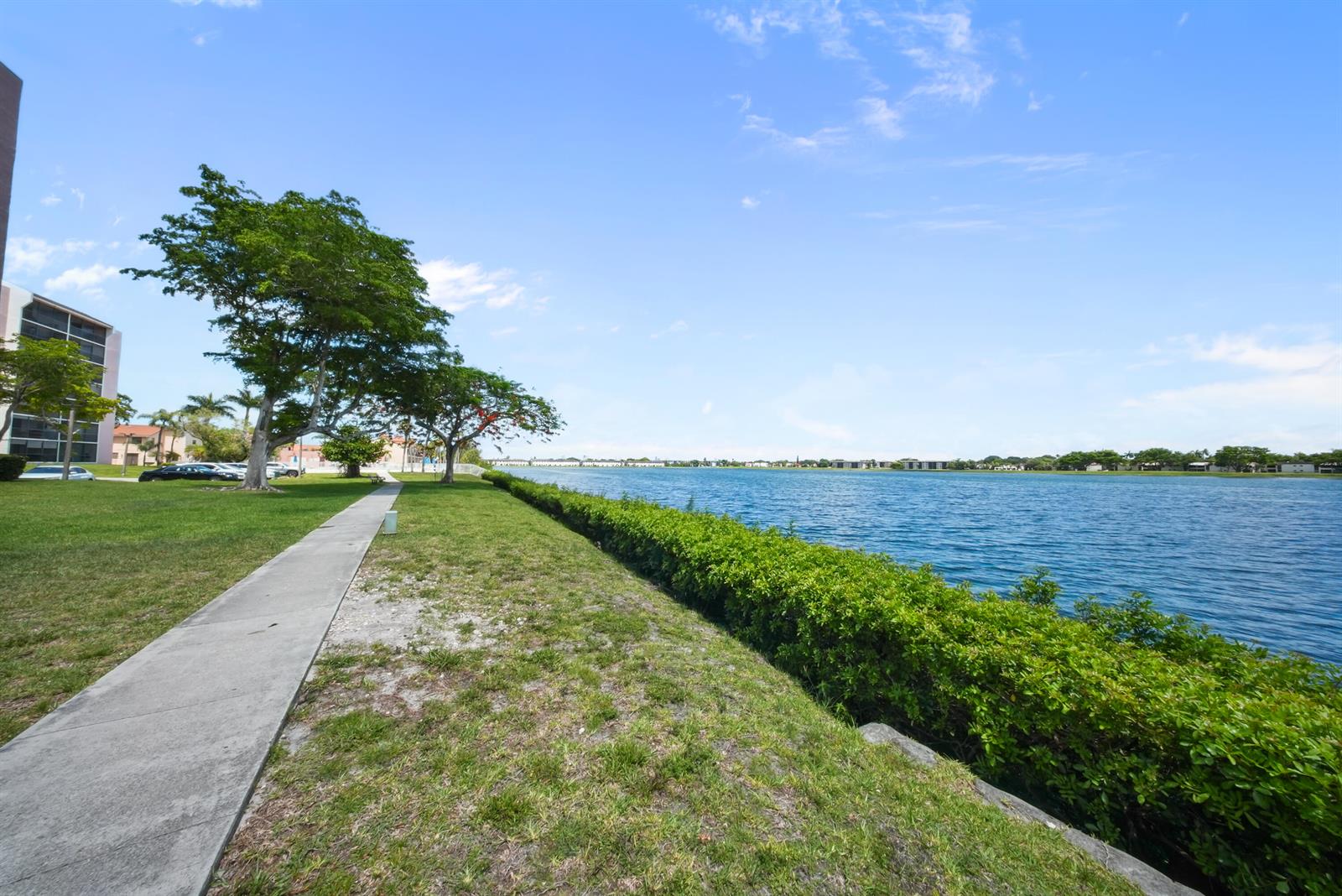 A concrete walkway runs along a grassy lawn beside a lake with a hedge border — 20860 San Simeon Way # 6, Miami FL 33179, MIAMI, FL