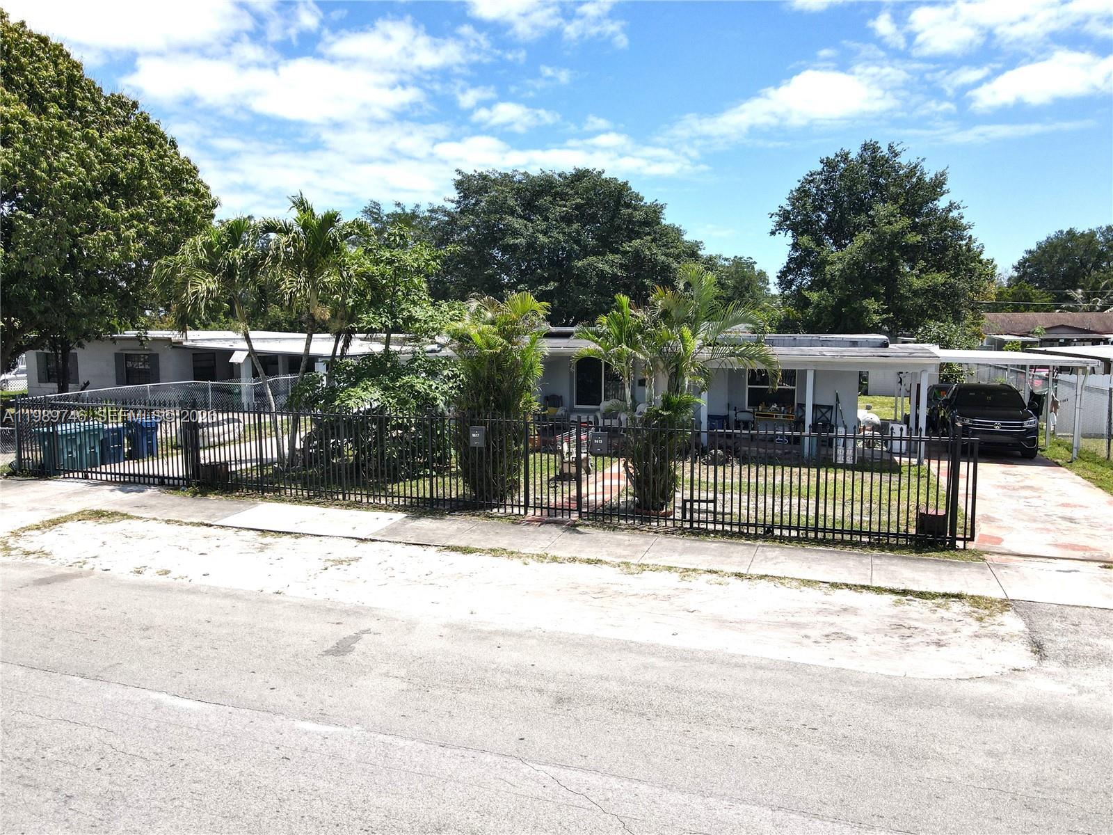 A single-story house with a fenced front yard and a carport sits under a blue sky — 9615 NW 26th Ave, Miami FL 33147, MIAMI, FL
