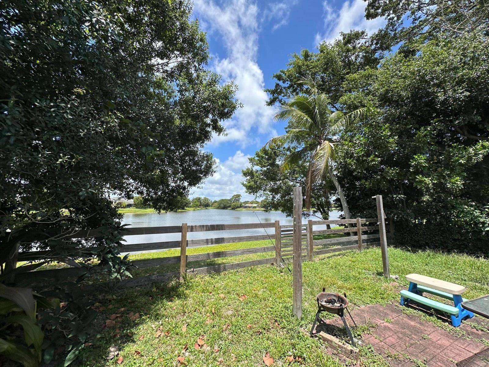 A backyard features a wooden fence, a small grill, and a bench overlooking a lake — 132 Timberline Drive, Jupiter FL 33458, JUPITER, FL