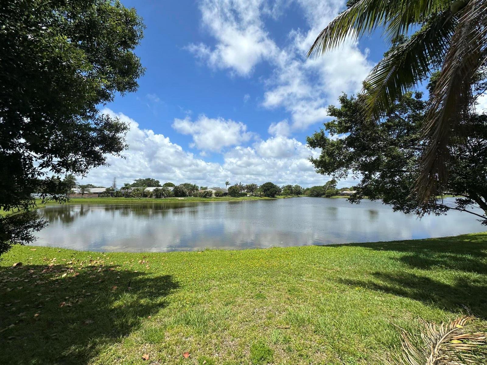 A grassy lawn overlooks a calm lake under a blue sky with scattered white clouds — 132 Timberline Drive, Jupiter FL 33458, JUPITER, FL