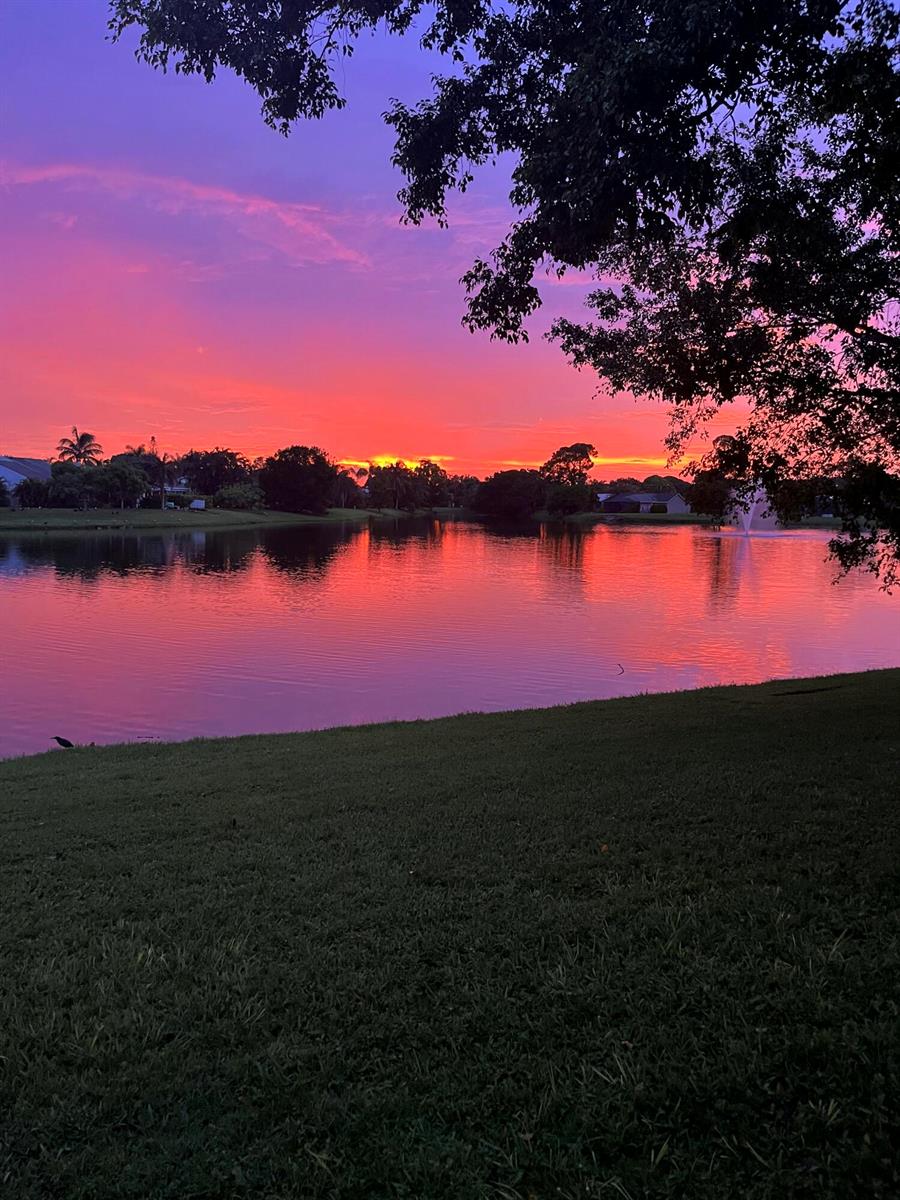 A vibrant sunset reflects over a calm lake with a fountain and surrounding grassy lawn — 132 Timberline Drive, Jupiter FL 33458, JUPITER, FL