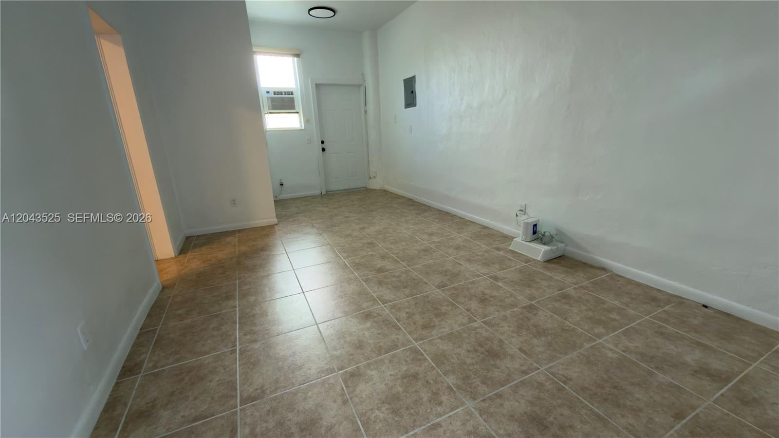 An empty room features white walls, tan tile flooring, a window, and a white door — 808 84th St # 812, Miami Beach FL 33141, MIAMI BEACH, FL