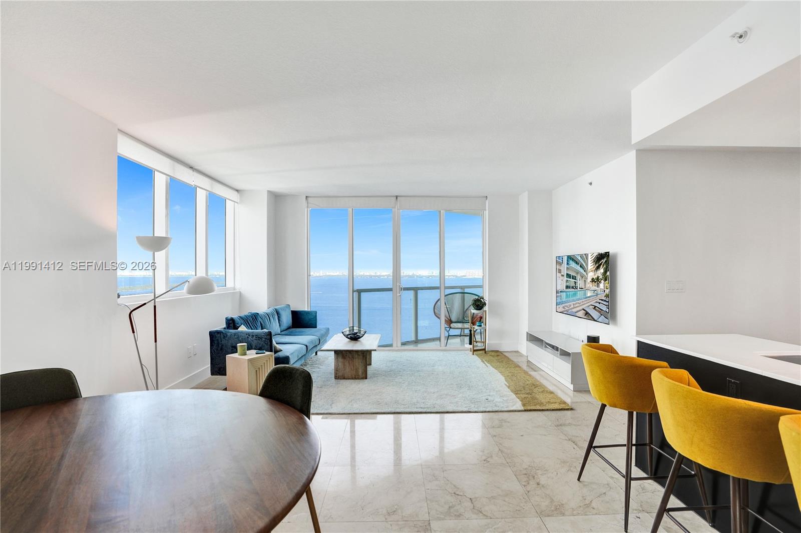 A modern living room features a blue sofa, yellow bar stools, and a balcony view — 665 NE 25th St # 2105, Miami FL 33137, MIAMI, FL