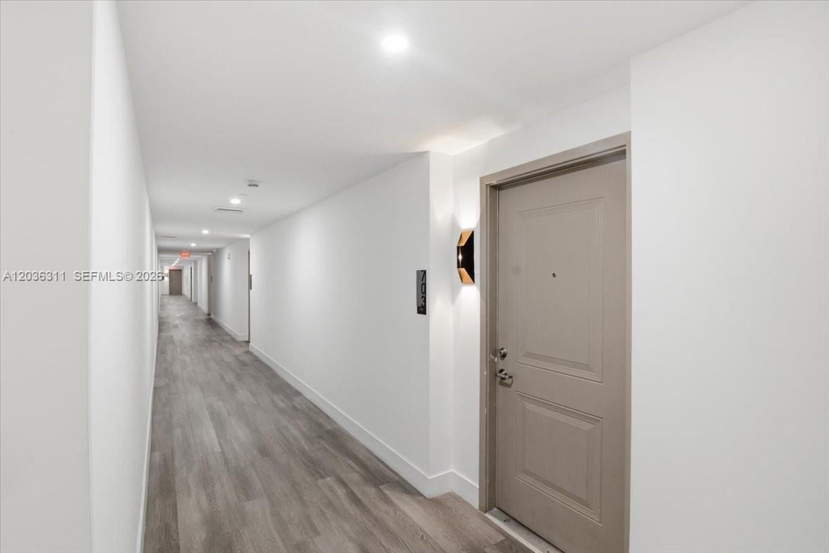A long, narrow apartment hallway features white walls, gray wood-look flooring, and a brown door — 1776 Polk St # 703, Hollywood FL 33020, HOLLYWOOD, FL