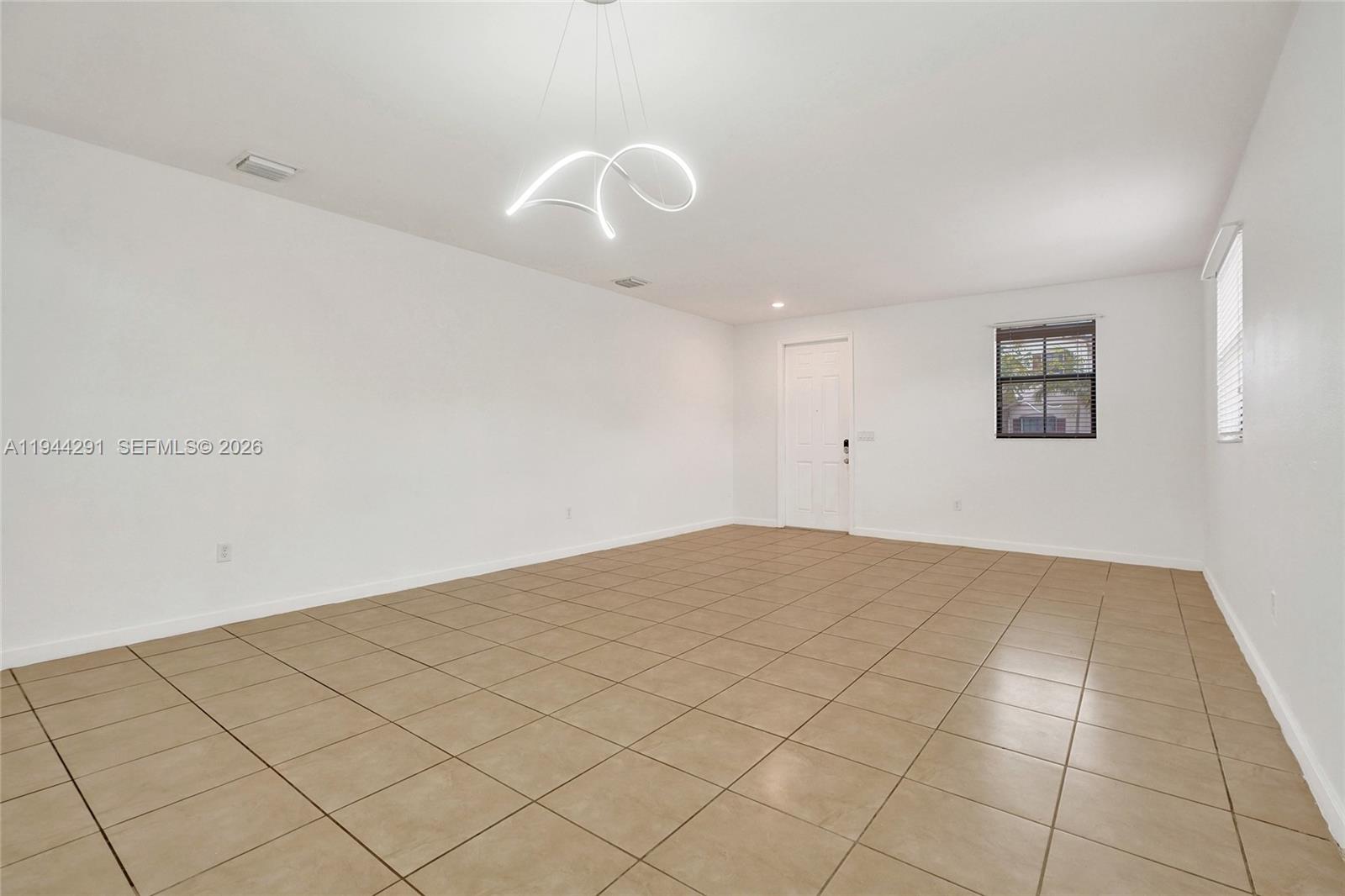 An empty room features white walls, beige tile flooring, and a modern sculptural light fixture — 3504 W 94th St, Hialeah FL 33018, HIALEAH, FL