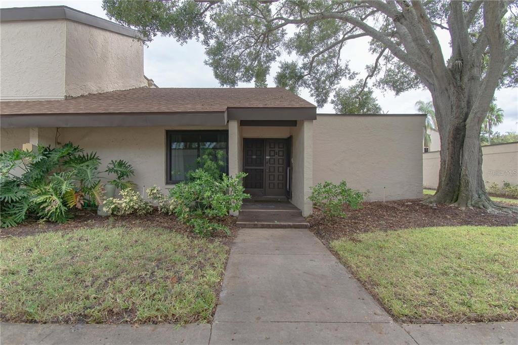 A concrete walkway leads to the front entrance of a tan house with a large tree — 2207 BELLEAIR ROAD UNIT B22, CLEARWATER, FL