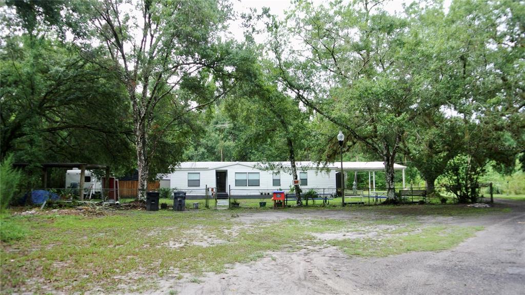A white single-wide mobile home sits on a dirt lot surrounded by large trees — 38122 CENTRAL AVENUE, ZEPHYRHILLS, FL