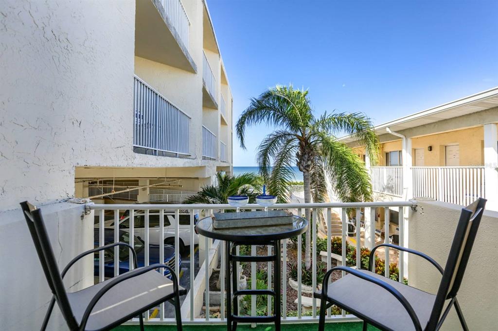 A balcony with two chairs and a small table overlooks a parking area and ocean — 3200 GULF BOULEVARD UNIT 201, BELLEAIR BEACH, FL