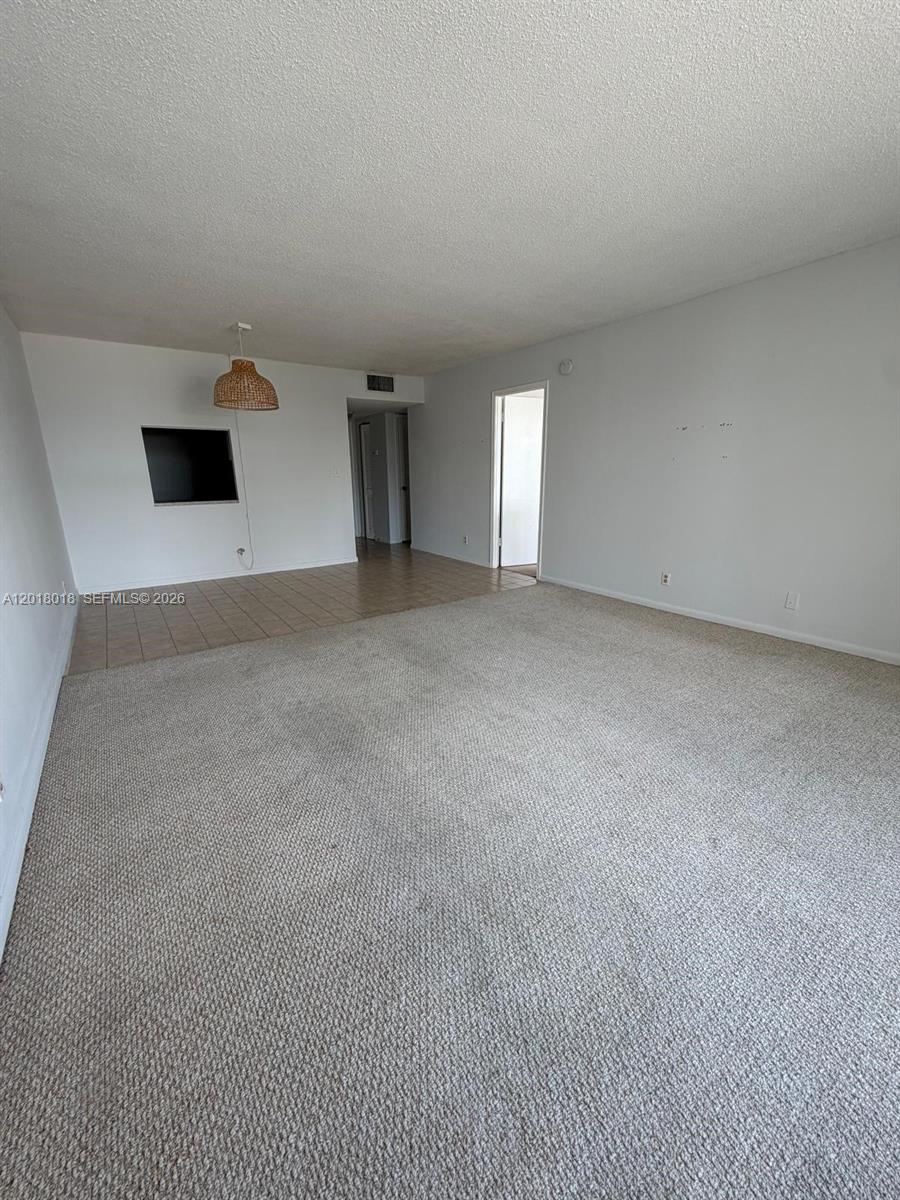 An empty living room features beige carpeting, a tiled dining area, and a hanging light — 1817 S Ocean Dr # 319, Hallandale Beach FL 33009, HALLANDALE BEACH, FL