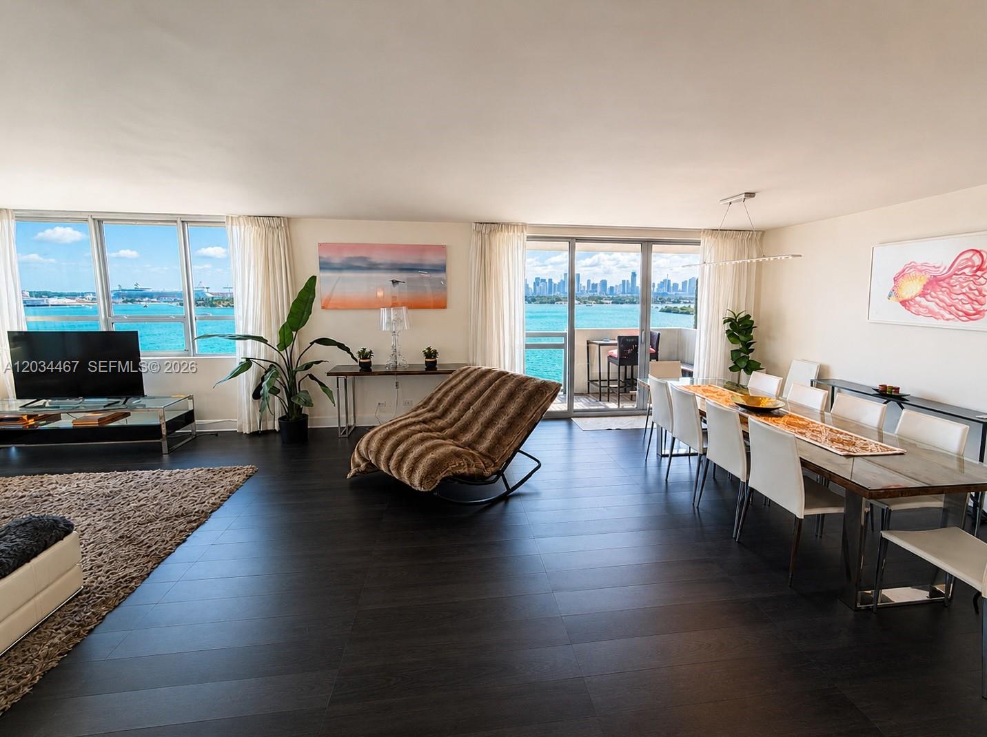 A spacious living and dining area features dark wood floors, floor-to-ceiling windows, and water views — MIAMI BEACH, FL