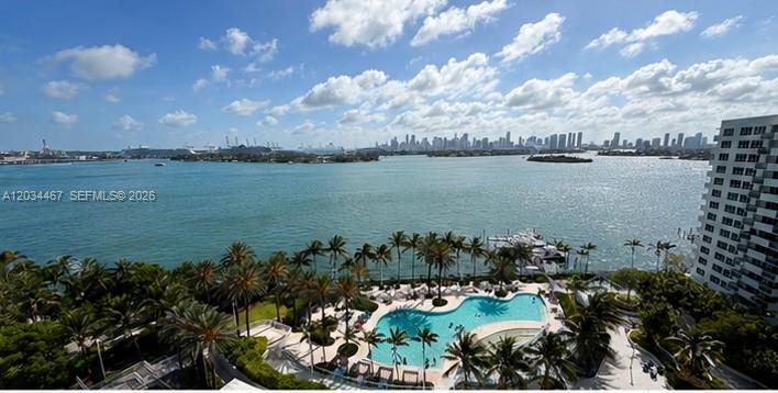 An elevated view of a swimming pool, palm trees, the bay, and a distant city skyline — MIAMI BEACH, FL
