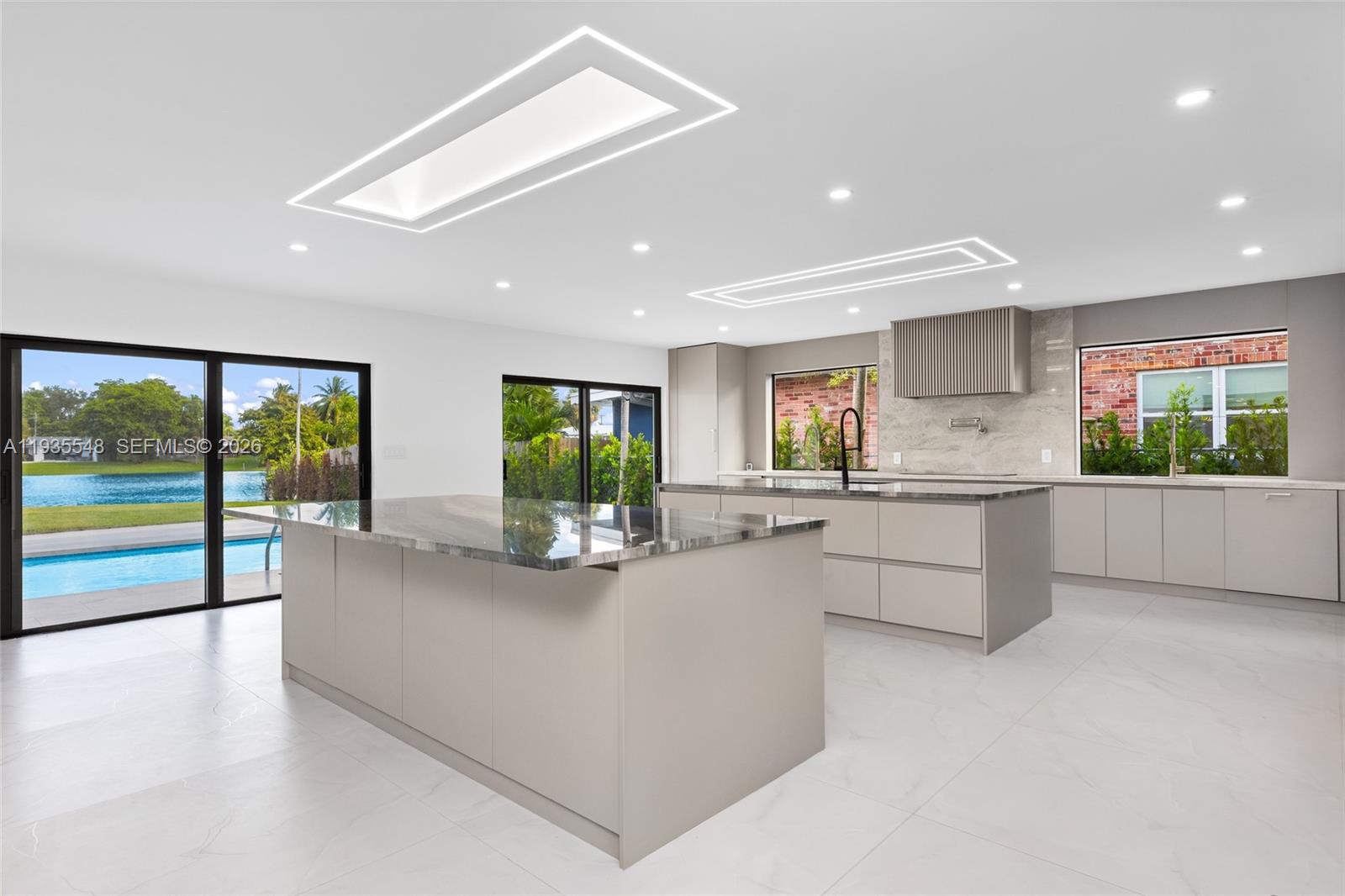 A modern kitchen features two large islands, light cabinetry, and sliding doors overlooking a pool — 18735 NE 18th Ave, Miami FL 33179, MIAMI, FL