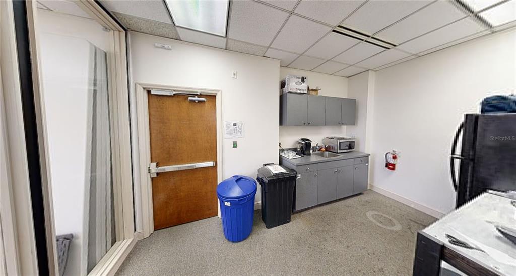 This office break room features gray cabinets, a small sink, two trash cans, and a door — 2815 NW 13TH STREET 2nd Floor, GAINESVILLE, FL