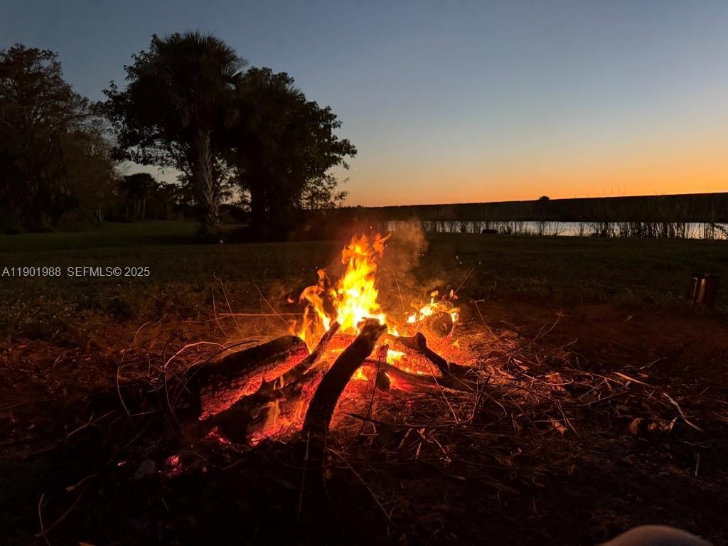 A campfire burns on the ground at dusk with trees and a lake in the background — 12600 SW Conners Highway, Okeechobee FL 34974, OKEECHOBEE, FL