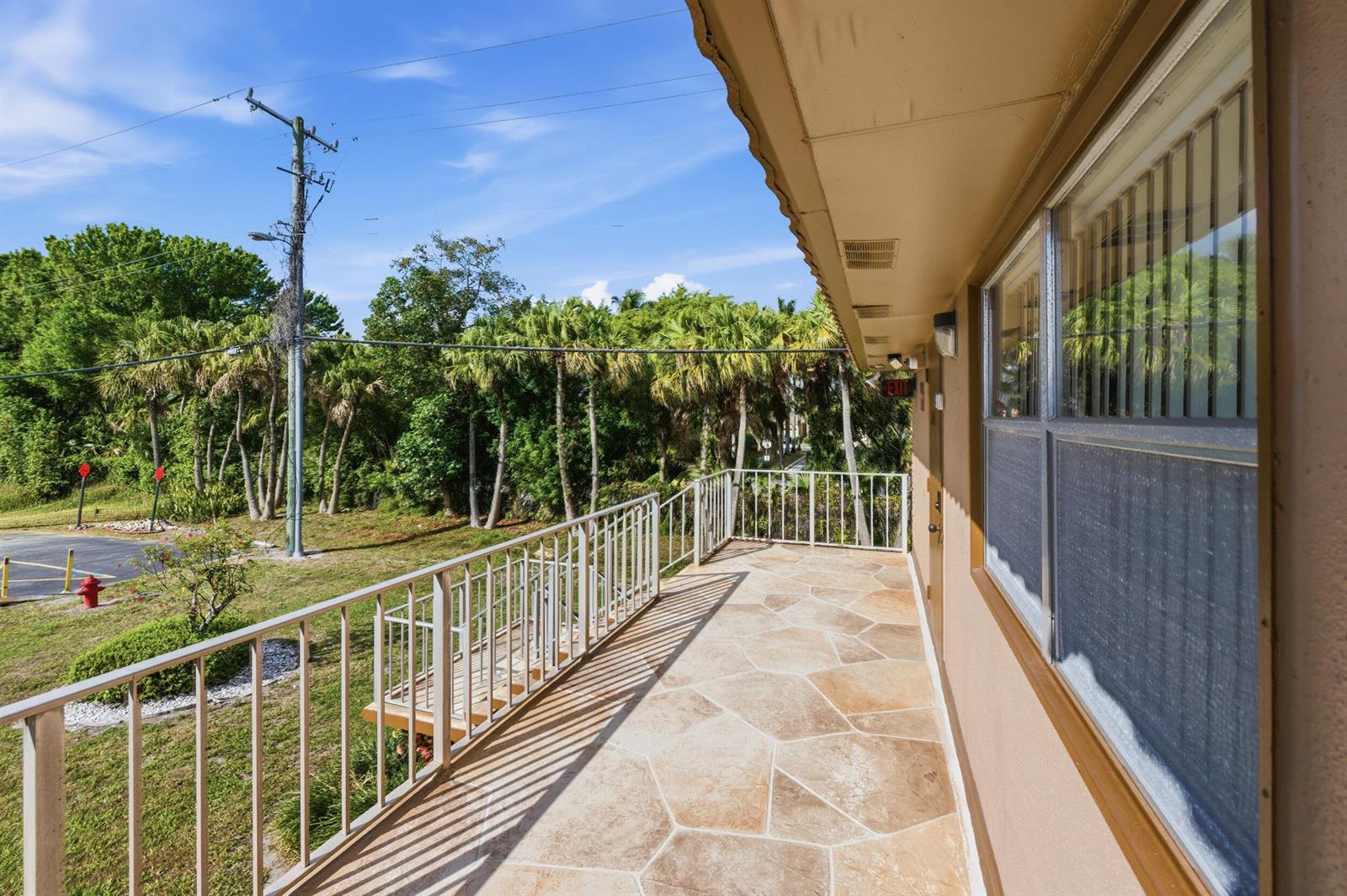 A balcony with stone-patterned flooring and a metal railing overlooks a grassy area with trees — 336 Brittany G, Delray Beach FL 33446, DELRAY BEACH, FL