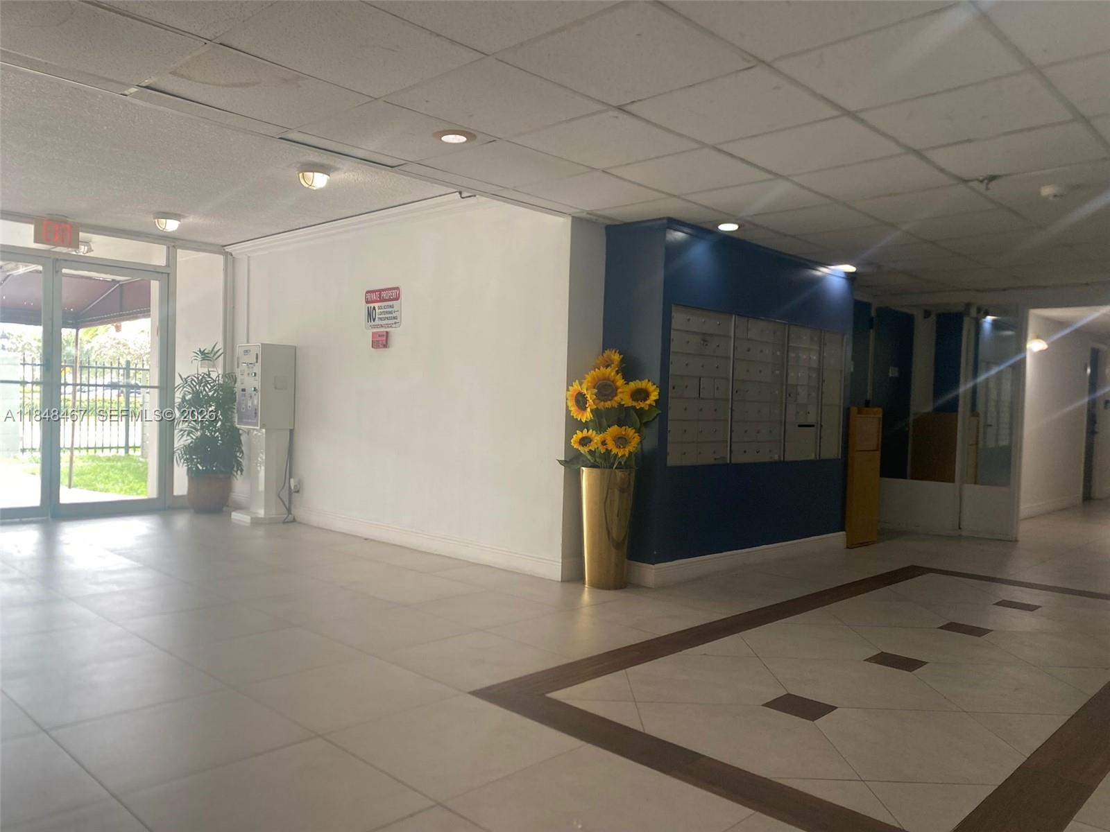 An indoor lobby features a tiled floor, mailboxes, a sunflower arrangement, and a glass entrance door — 16450 NW 2nd Ave # 404, Miami FL 33169, MIAMI, FL