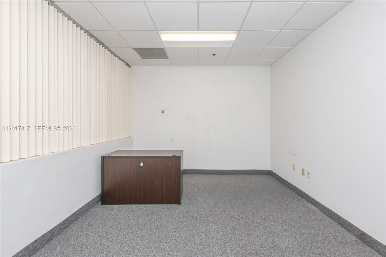 A small, empty office features gray carpeting, white walls, a drop ceiling, and a dark desk — 1900 Glades Rd # 280, Boca Raton FL 33431, BOCA RATON, FL