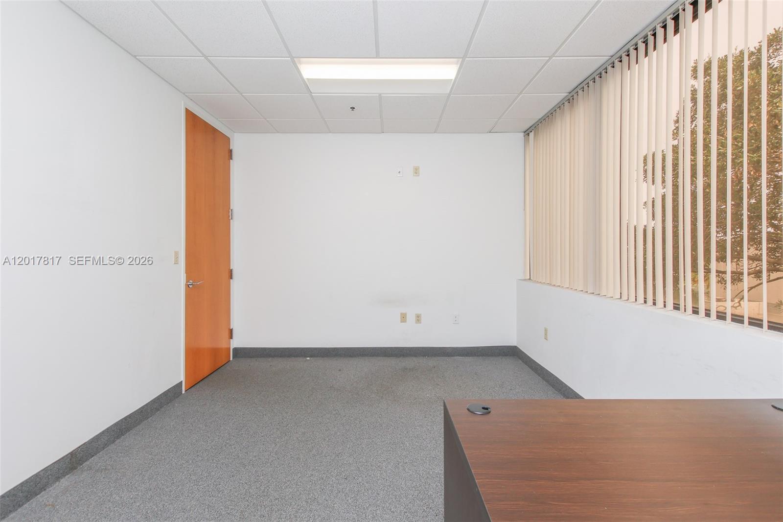 An empty office features white walls, gray carpet, a wooden door, and vertical window blinds — 1900 Glades Rd # 280, Boca Raton FL 33431, BOCA RATON, FL
