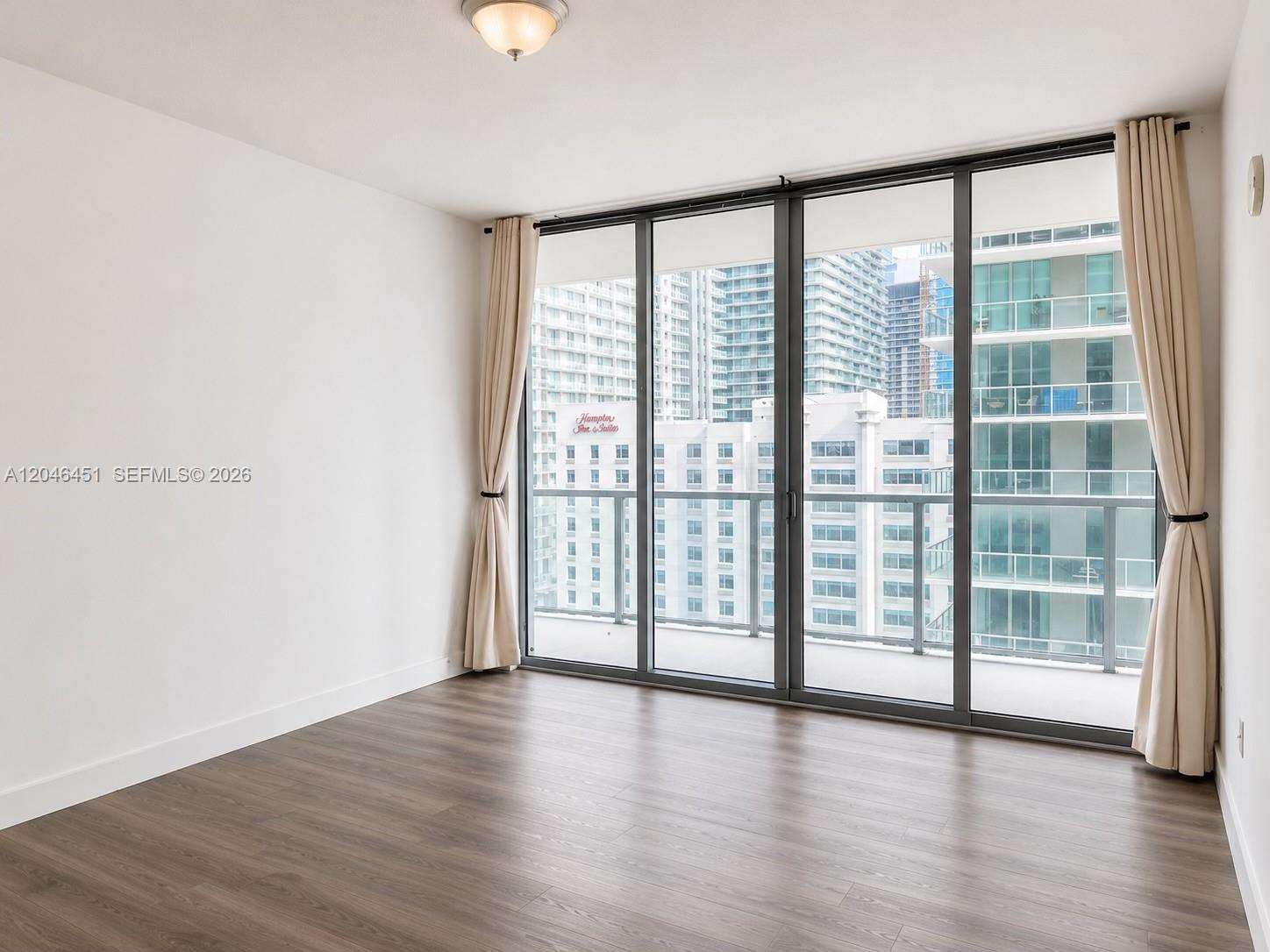 An empty room features wood-look flooring, white walls, and floor-to-ceiling windows overlooking city buildings — 1111 SW 1st Ave, Miami FL 33130, MIAMI, FL