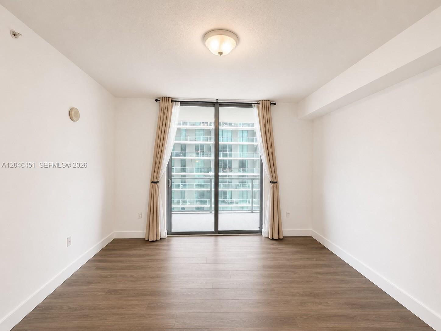 An empty room features wood-look flooring, white walls, a ceiling light, and a sliding glass door — 1111 SW 1st Ave, Miami FL 33130, MIAMI, FL