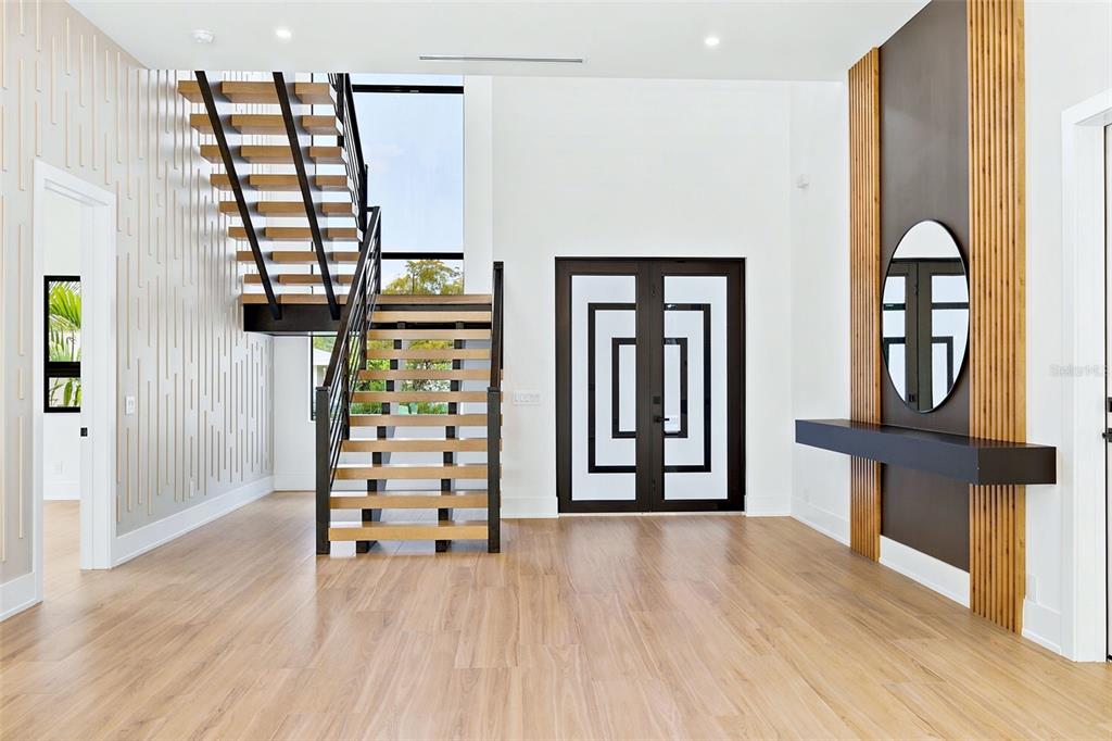 A modern entryway features light wood flooring, a floating staircase, and a decorative wood-slatted accent wall — 1870 JASMINE DRIVE, SARASOTA, FL