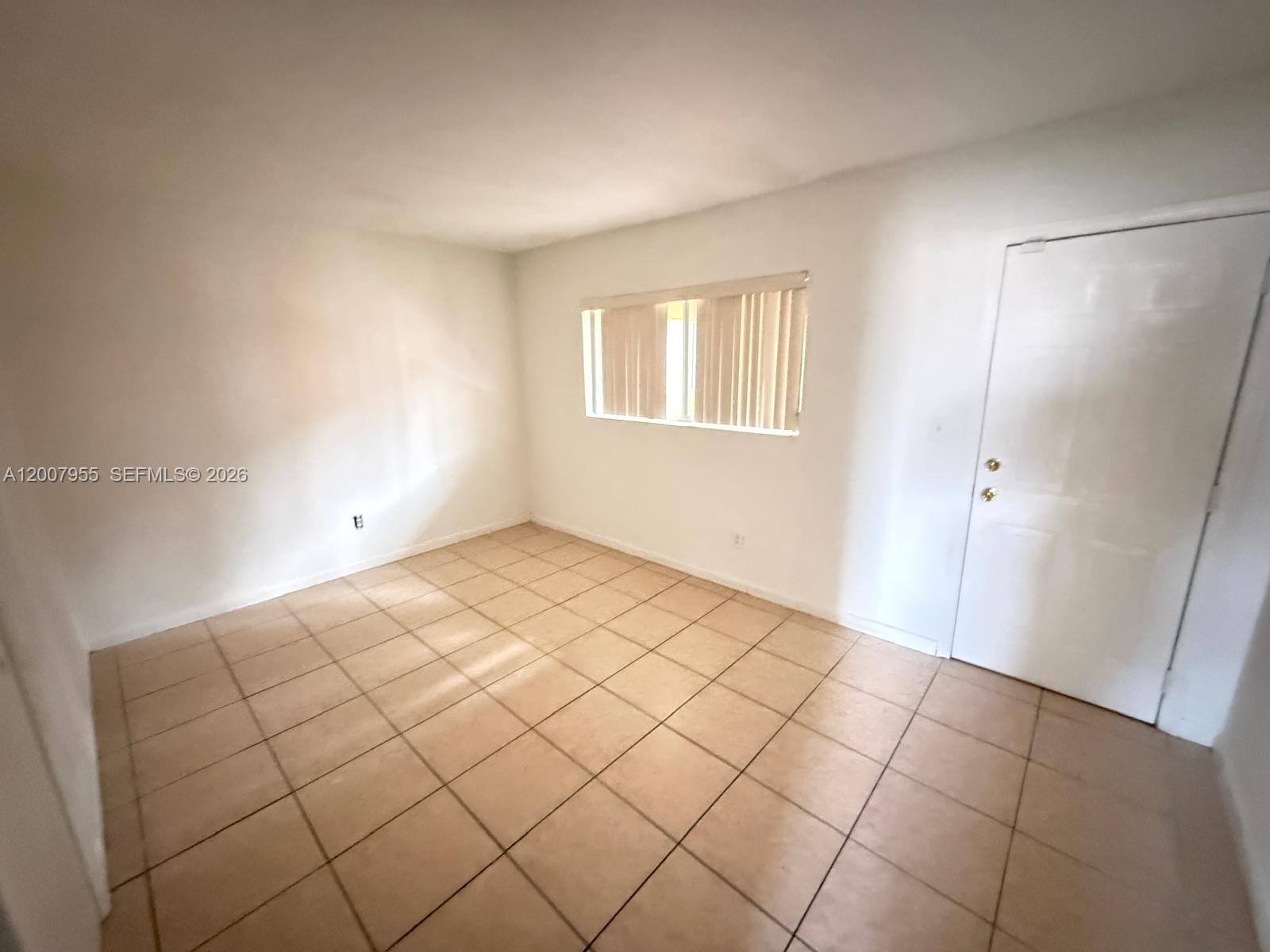An empty room features white walls, beige tile flooring, a window with blinds, and a door — 2330 NW 11th St # 33, Miami FL 33125, MIAMI, FL