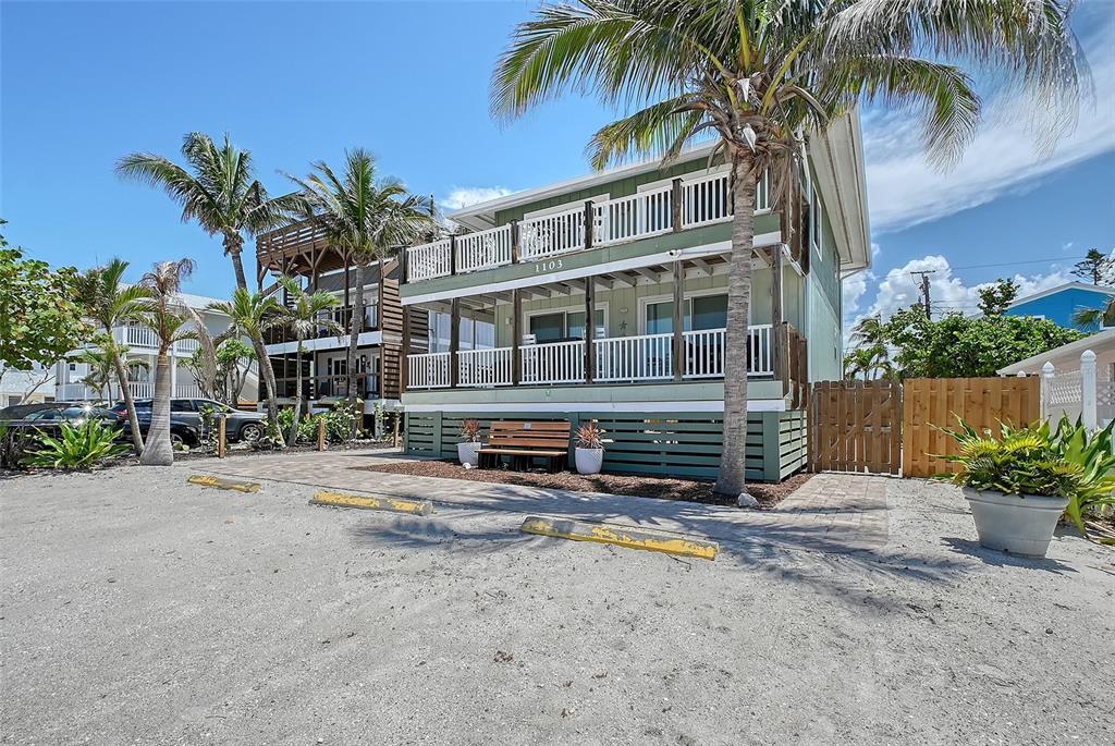 A three-story green house with white railings and palm trees stands above a gravel parking area — 1103 SHORE VIEW DRIVE, ENGLEWOOD, FL