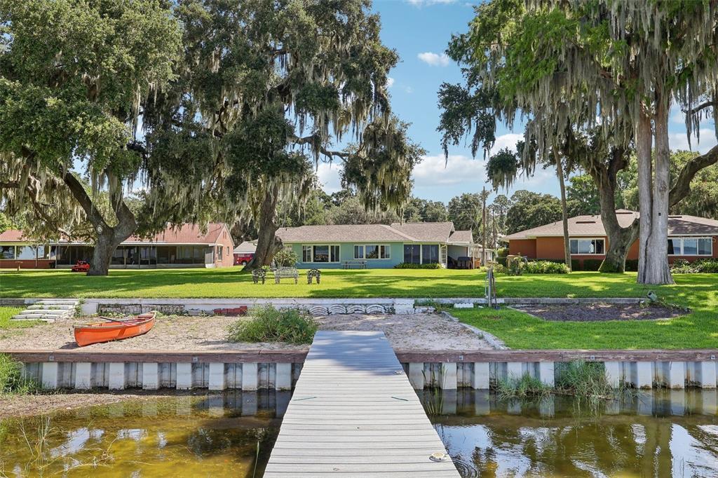 A wooden dock extends toward a grassy lawn with three houses and large oak trees — 31929 ELIZABETH LANE, TAVARES, FL
