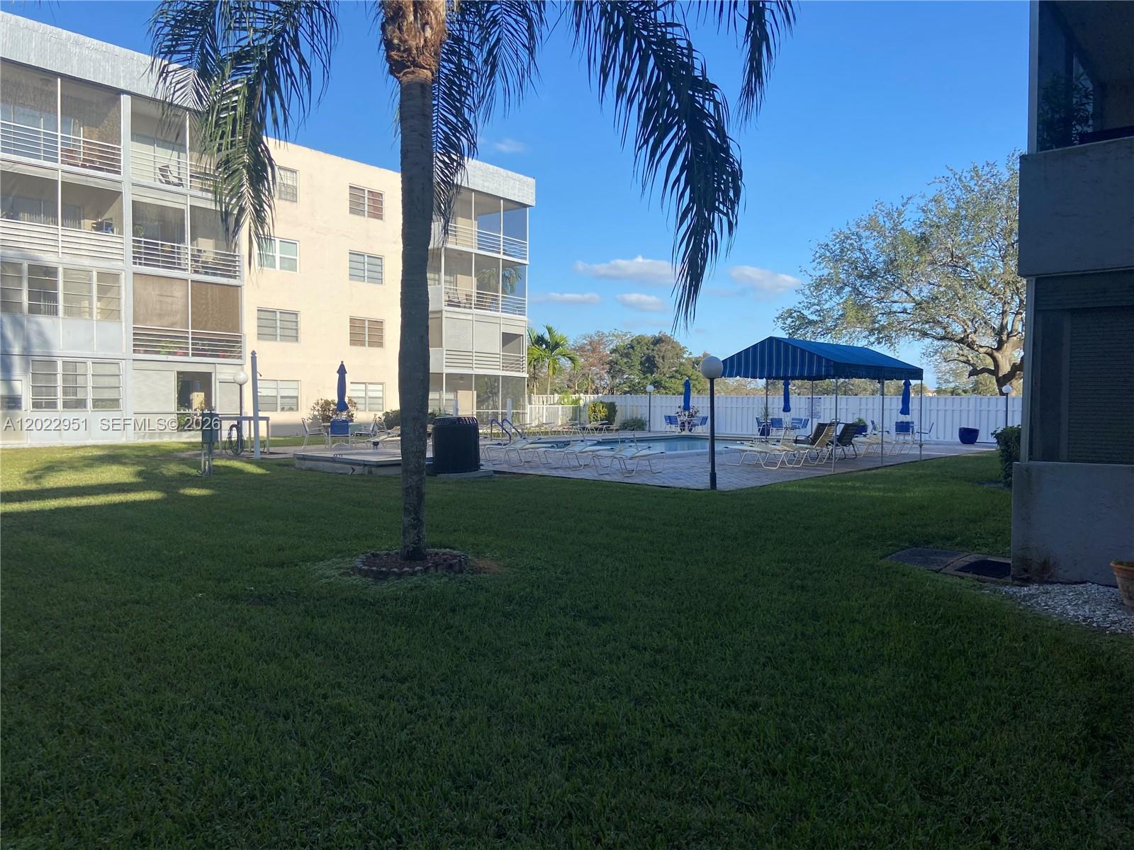 A swimming pool area with lounge chairs and a blue canopy sits beside an apartment building — 7502 NW 30th Pl # 417, Sunrise FL 33313, SUNRISE, FL