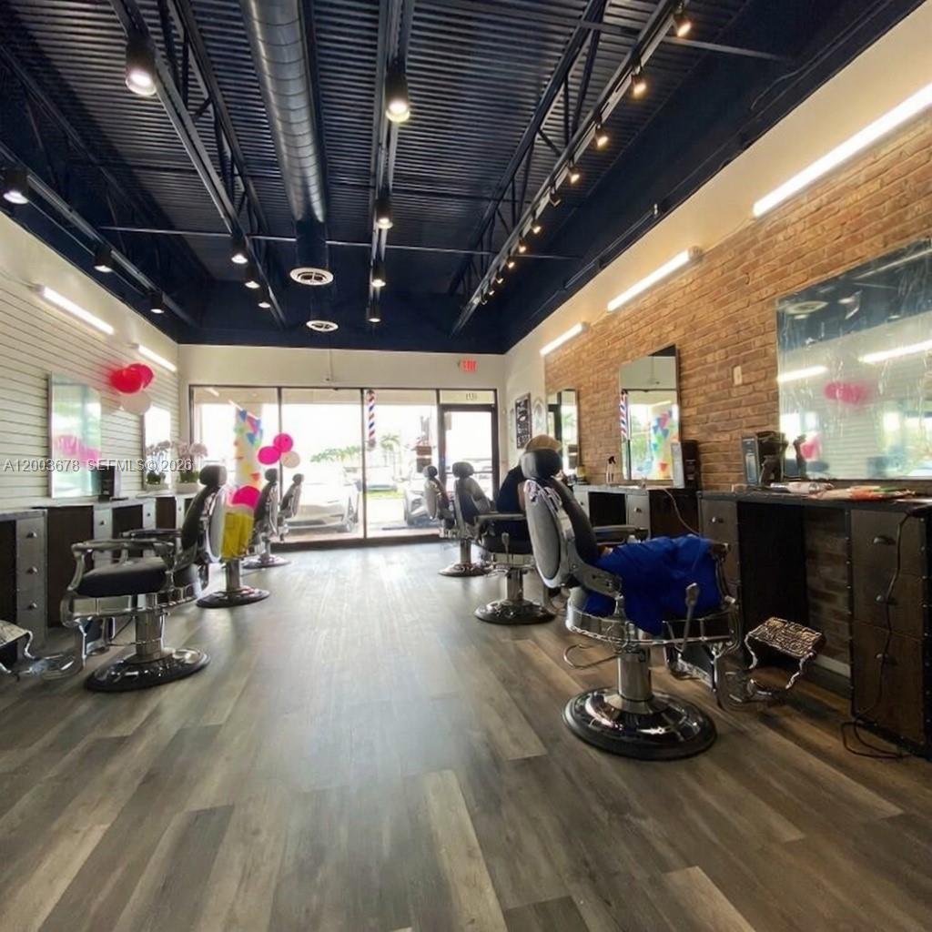 A barbershop interior features multiple styling chairs, mirrors, brick walls, and a dark ceiling — AVENTURA, FL