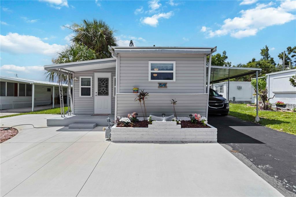 A light gray manufactured home with a white door, carport, and a small front garden — 1100 UNIVERSITY PARKWAY UNIT 54, SARASOTA, FL