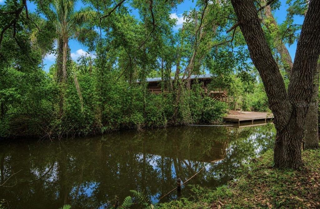 A wooden house sits nestled among lush trees beside a calm, reflective canal — 707 GEORGE STREET S, TARPON SPRINGS, FL