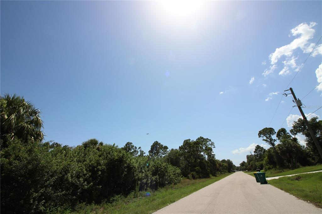A paved road runs alongside a dense, wooded lot under a bright, sunny sky — 26351 BROOKS ROAD, PUNTA GORDA, FL