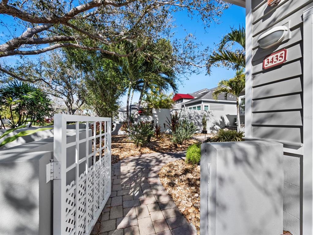 A white gate opens onto a stone walkway leading toward a house with tropical landscaping — 3435 WINDING OAKS DRIVE UNIT 18, LONGBOAT KEY, FL
